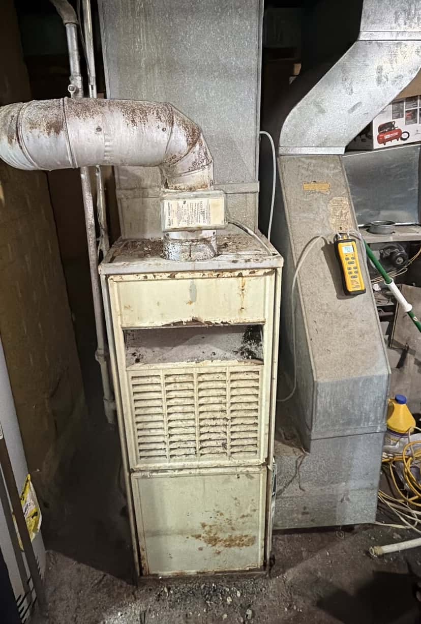 Old, rusted gas furnace with a corroded metal vent pipe in an unfinished basement